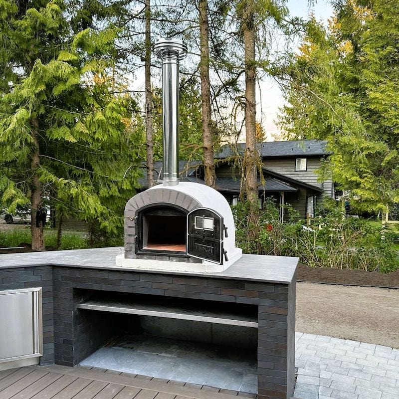 Lume Alto Premium Oven on  an outdoor kitchen countertop with black brick finish and trees and a house behind