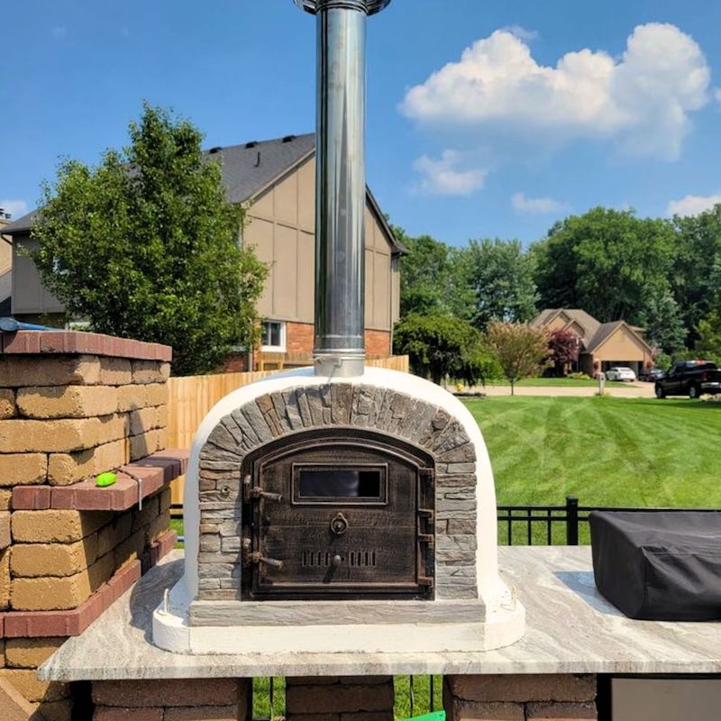 Ventura Preto Pizza Oven with Chimney Extension, set on a countertop in a suburban backyard with green grass, trees, and houses in the background