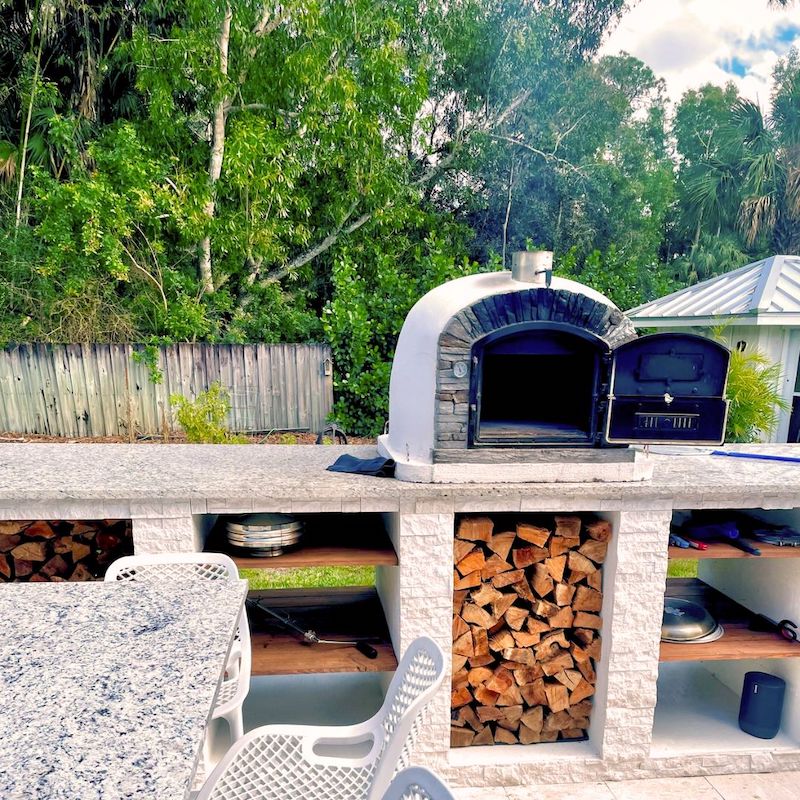 Ventura Preto Outdoor Pizza Oven cooling down, set on an outdoor kitchen countertop with firewood stacked underneath