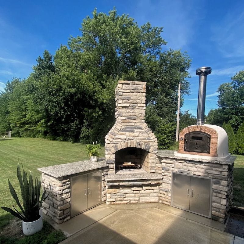 Ventura Sierra Wood Fired Brick Oven sitting on a granite outdoor kitchen countertop next to a fireplace in a garden with trees in the background