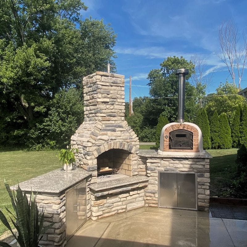 Ventura Sierra Wood Fired Brick Oven sitting on a granite outdoor kitchen countertop next to a fireplace in a garden